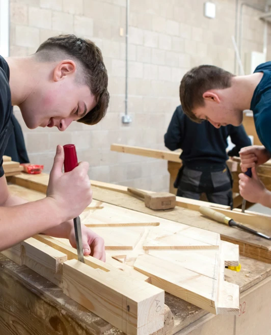 Two joinery students concentrating deeply, using chisels to precisely shape wooden components on a workbench during a practical woodworking class. Two joinery students concentrating deeply, using chisels to precisely shape wooden components on a workbench during a practical woodworking class.